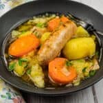 A black bowl of Dublin Coddle filled with sausage, carrots, potatoes, and vegetables in broth, with a fork resting on the edge.