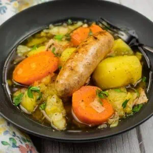 A black bowl of Dublin Coddle filled with sausage, carrots, potatoes, and vegetables in broth, with a fork resting on the edge.