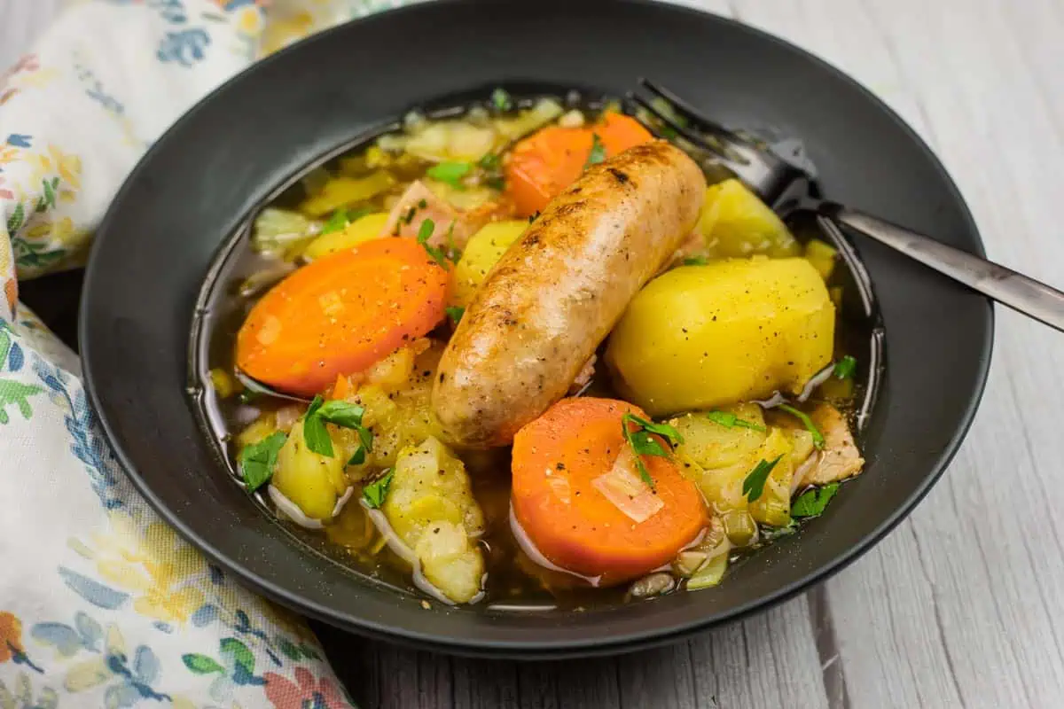 A black bowl of Dublin Coddle filled with sausage, carrots, potatoes, and vegetables in broth, with a fork resting on the edge.