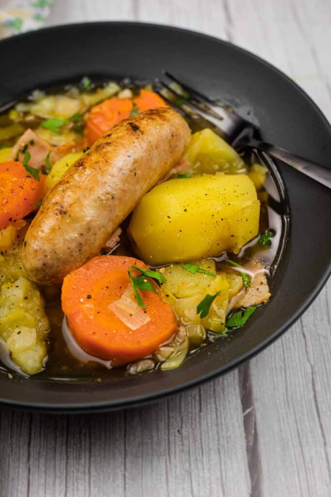 A black bowl containing sausage, potatoes, carrots, and cabbage in broth, garnished with parsley, with a fork on the side.