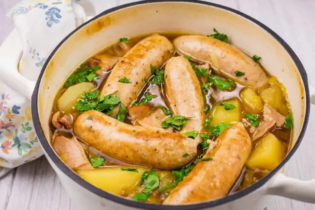 A pot filled with sausages, potatoes, and herbs in broth, garnished with chopped parsley, on a light wooden surface.