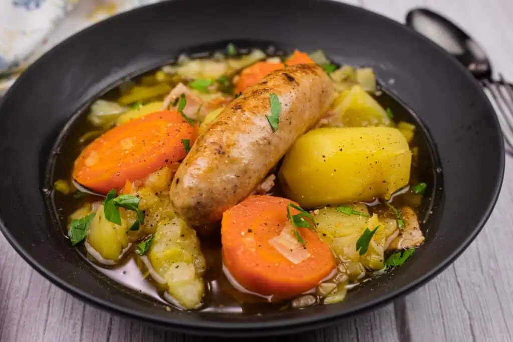 A bowl of Dublin Coddle with a sausage, sliced carrots, potatoes, and herbs in a dark broth, served in a black dish.