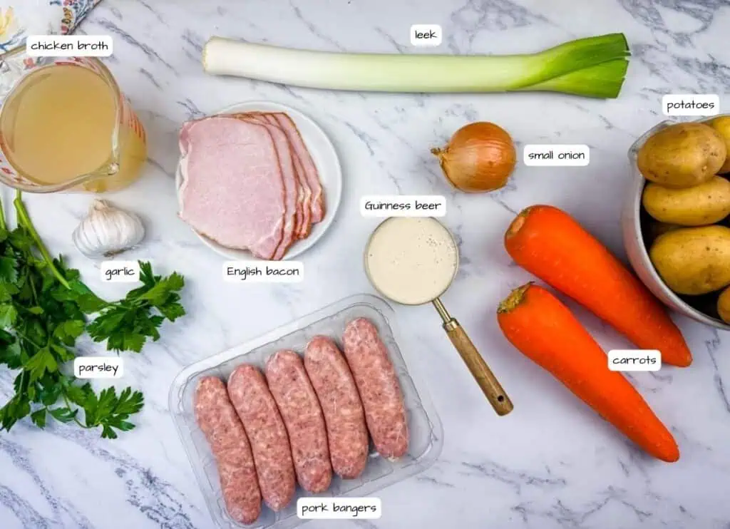 Ingredients for a meal laid out on a marble surface, including chicken broth, leek, potatoes, carrots, onion, garlic, parsley, English bacon, Guinness beer, and pork sausages.