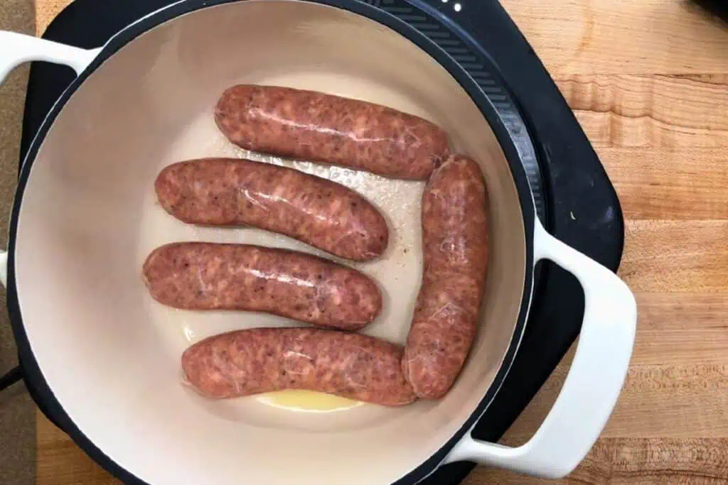Five raw sausages are arranged in a white pot on a stovetop, with a small amount of oil visible at the bottom.