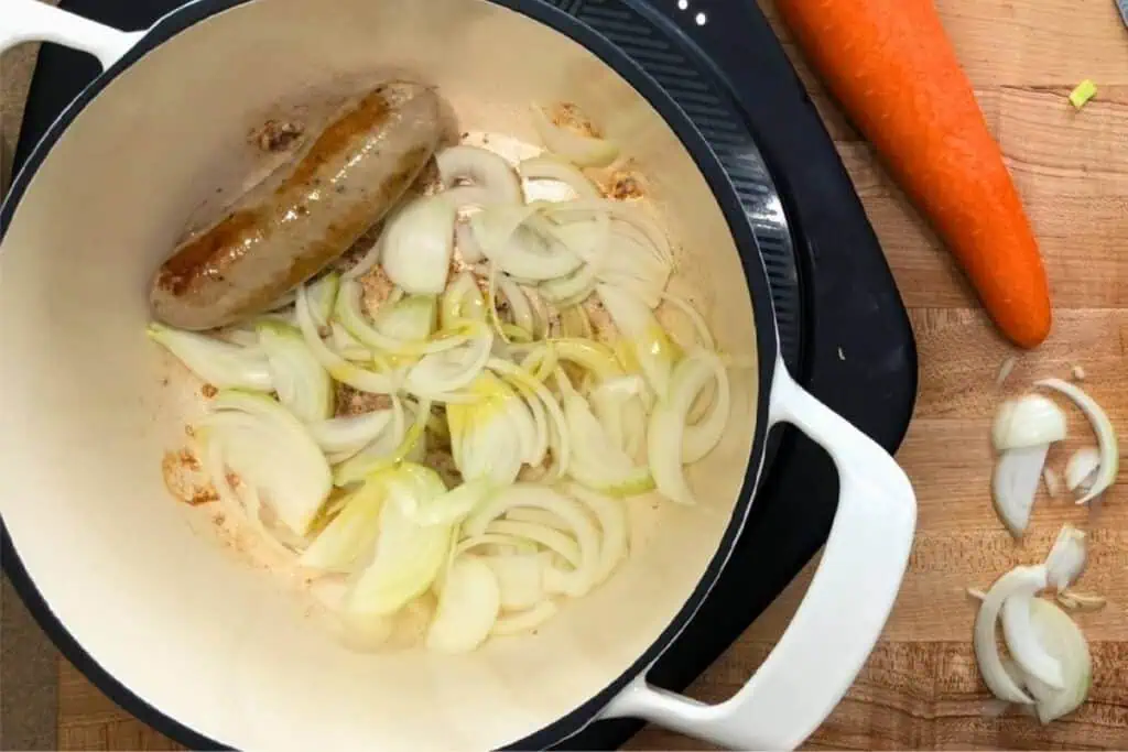 A white pot with sliced onions and a browned sausage inside, placed on a cooktop; a whole carrot and more sliced onions are on a wooden cutting board beside it.