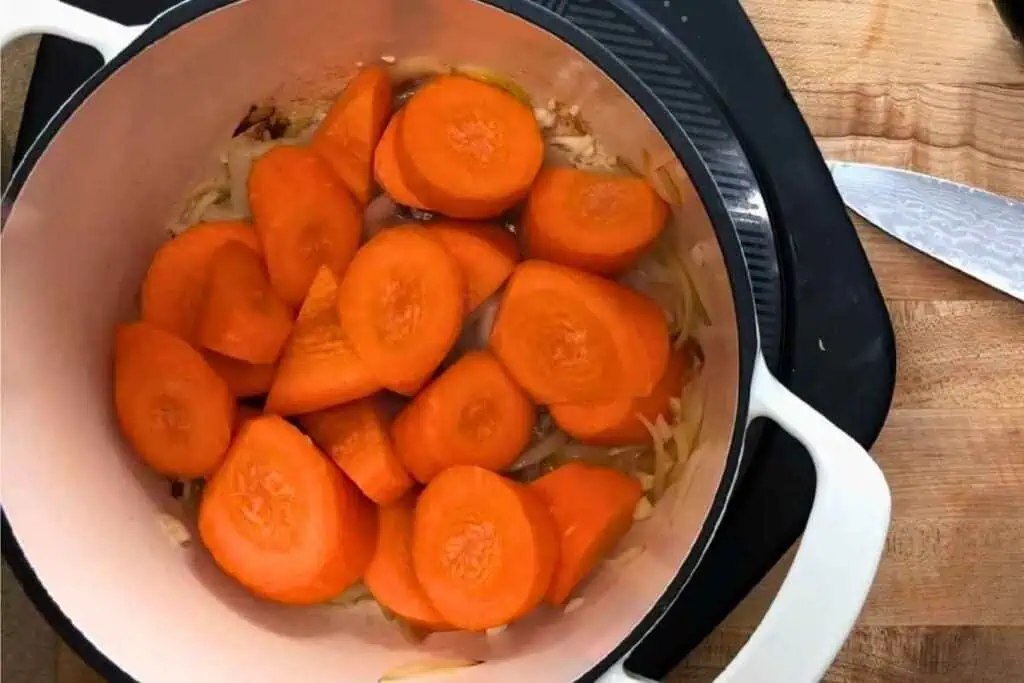 Sliced carrots and onions in a white pot on a stovetop next to a kitchen knife and wooden cutting board.