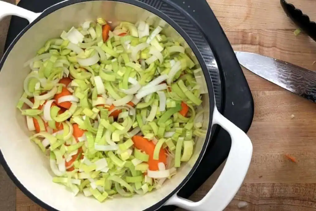 A white pot on a stovetop contains chopped leeks, onions, and carrots. A knife and cutting board with vegetable scraps are beside the pot.