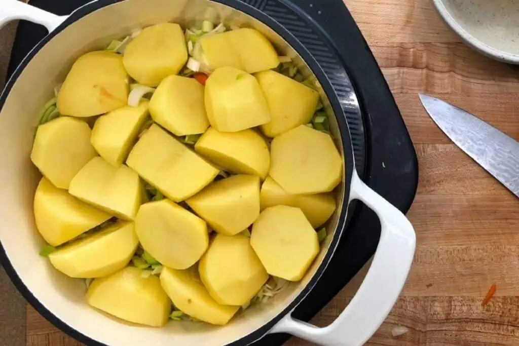 A white pot filled with peeled and sliced potatoes on top of chopped vegetables, placed on a stovetop beside a knife and a bowl on a wooden counter.