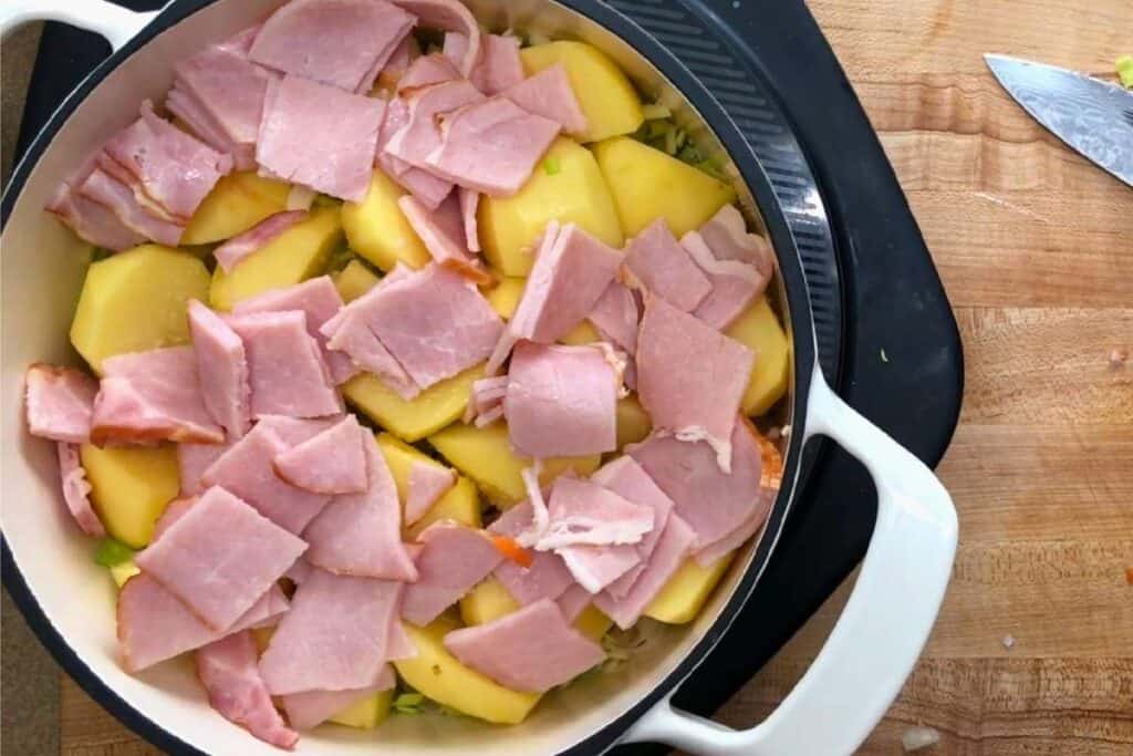 A white pot filled with sliced potatoes and pieces of ham sits on a black stovetop beside a wooden cutting board with a knife.