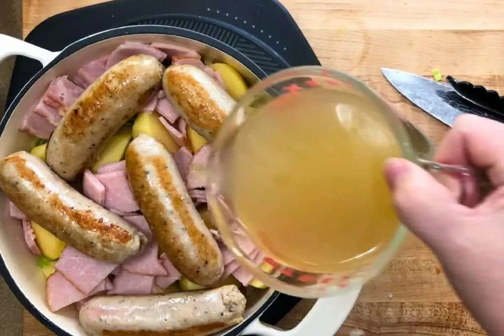 A hand pours broth from a measuring cup over sausages, ham, and sliced potatoes in a white pot on a wooden countertop.