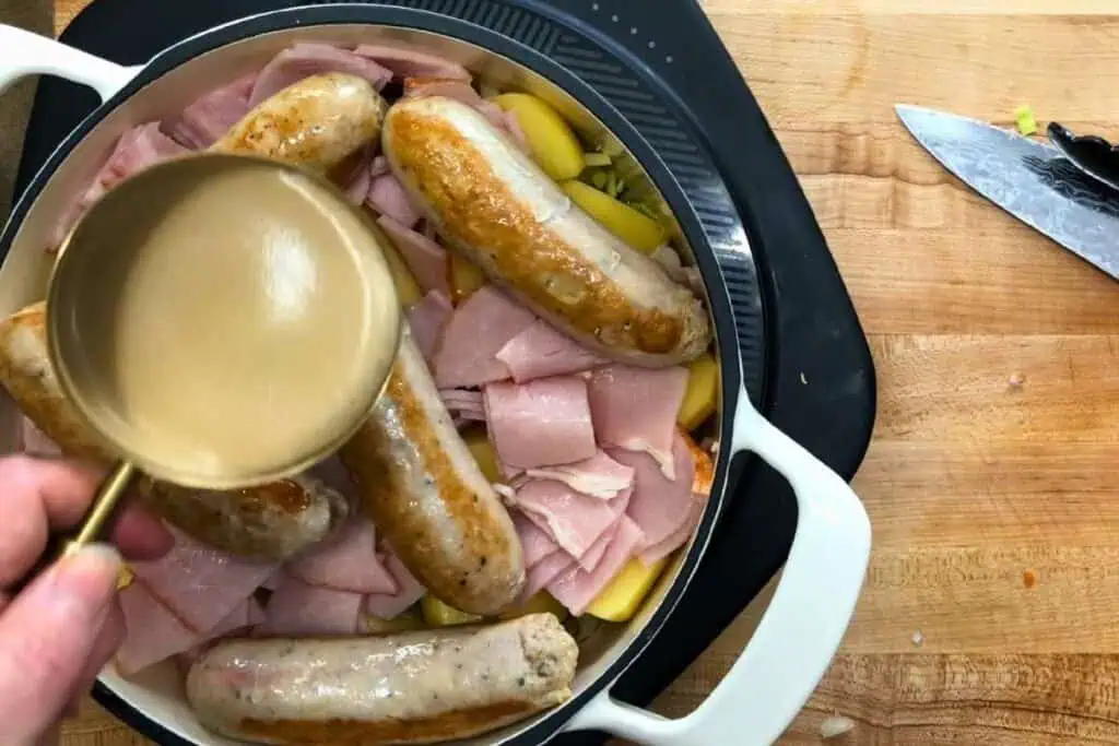 A hand pours liquid from a measuring cup over sausages and sliced ham arranged in a white Dutch oven on a wooden countertop.