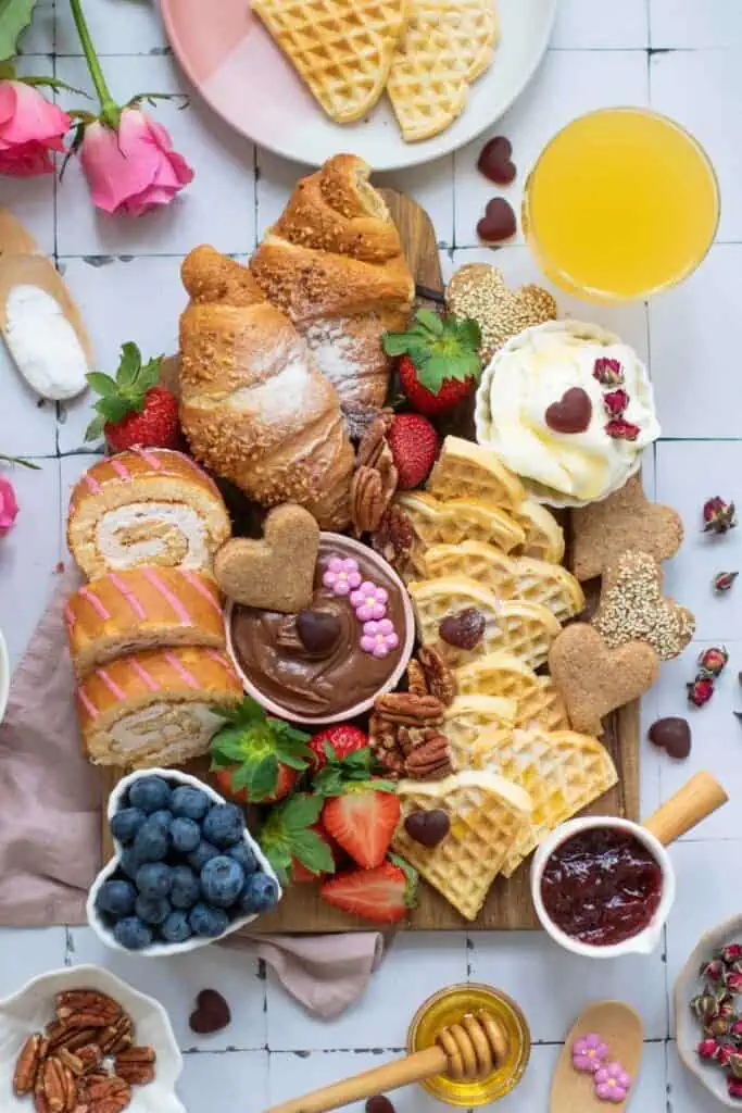 A breakfast board with croissants, waffles, cake rolls, fruit, nuts, jam, honey, chocolate spread, and a glass of orange juice, decorated with heart-shaped treats and flowers.