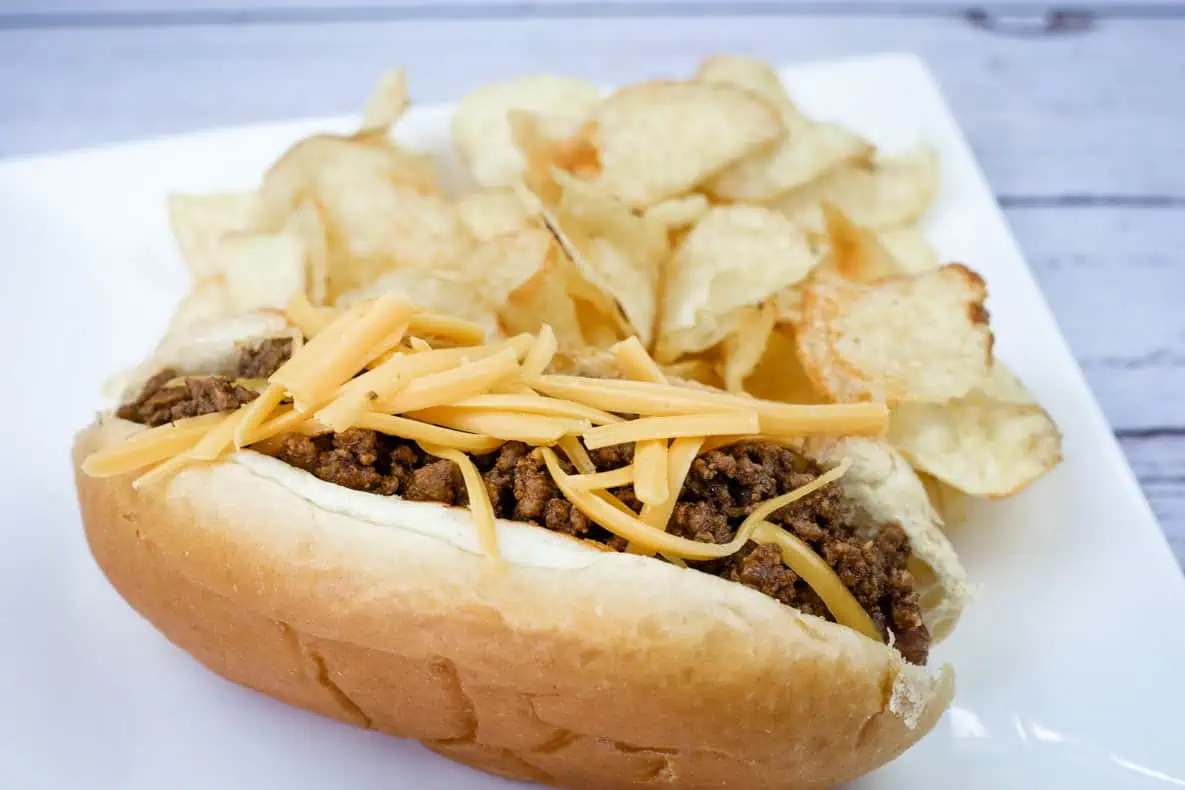 A hot dog chili topped with shredded cheddar, served with potato chips on a white plate.