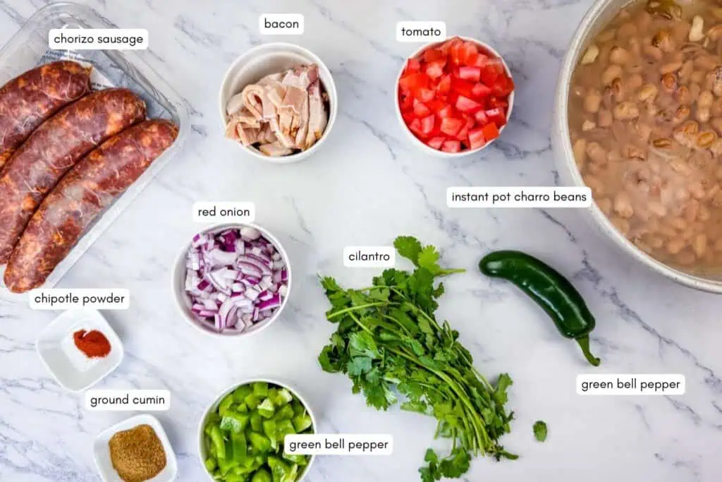 Chorizo sausages, chipotle powder, ground cumin, chopped red onion, green bell pepper, cilantro, bacon, diced tomato, green bell pepper, and a pot of charro beans on a marble surface.