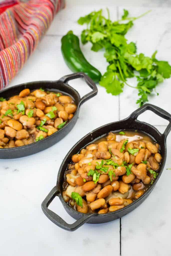 Two oval cast iron dishes filled with cooked pinto beans, garnished with chopped cilantro, sit on a white surface next to fresh cilantro, a jalape&ntilde;o, and a striped cloth.