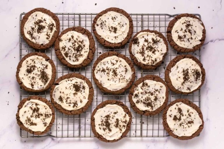 Twelve chocolate cookies with white icing and chocolate crumbs are arranged on a wire cooling rack over a marble surface.