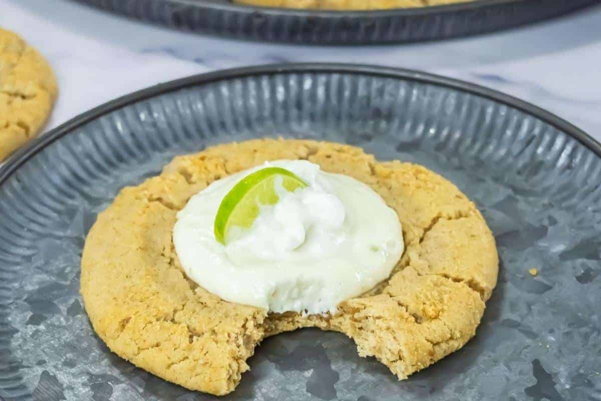 A large Copycat Crumbl Key Lime Cookies with a bite taken out, topped with white cream and a lime wedge, served on a gray plate.