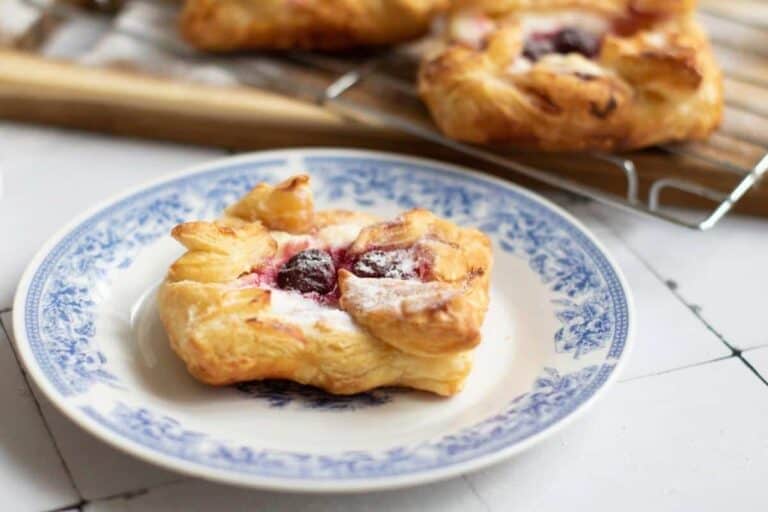 A cherry danish pastry with powdered sugar on a decorative blue and white plate, with more pastries cooling on a rack in the background.