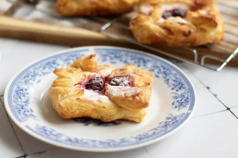 A cherry danish pastry with powdered sugar on a decorative blue and white plate, with more pastries cooling on a rack in the background.