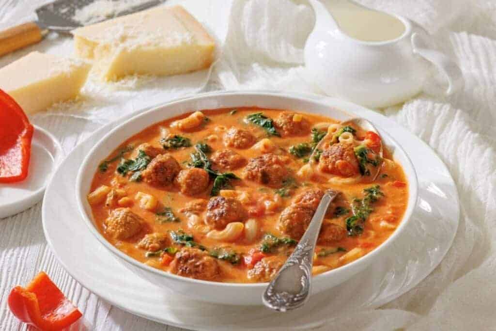A bowl of meatball soup with pasta, spinach, and tomato broth, served with a spoon.