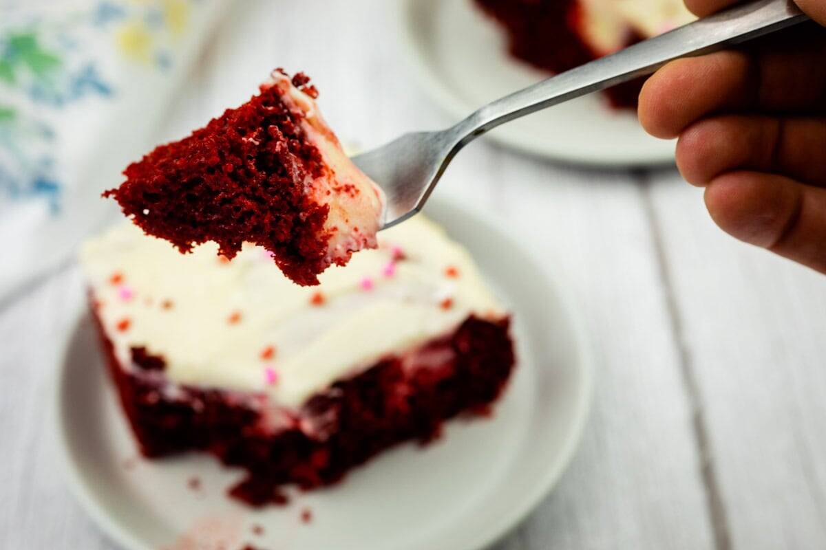A hand holds a fork with a bite of red velvet poke cake topped with white frosting above a plate containing the rest of the cake slice.