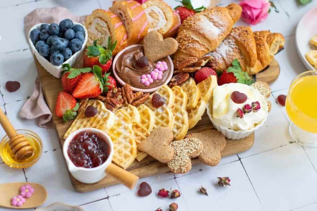 A wooden board with waffles, croissants, swiss rolls, strawberries, blueberries, nuts, chocolate spread, whipped cream, jam, honey, and cookies, with a glass of orange juice nearby.