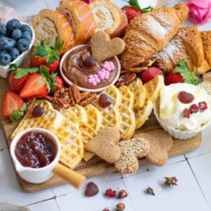 A Valentine's breakfast platter with waffles, croissants, strawberries, blueberries, jam, whipped cream, chocolate spread, cookies, and orange juice on a wooden board.