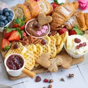 A Valentine's breakfast platter with waffles, croissants, strawberries, blueberries, jam, whipped cream, chocolate spread, cookies, and orange juice on a wooden board.