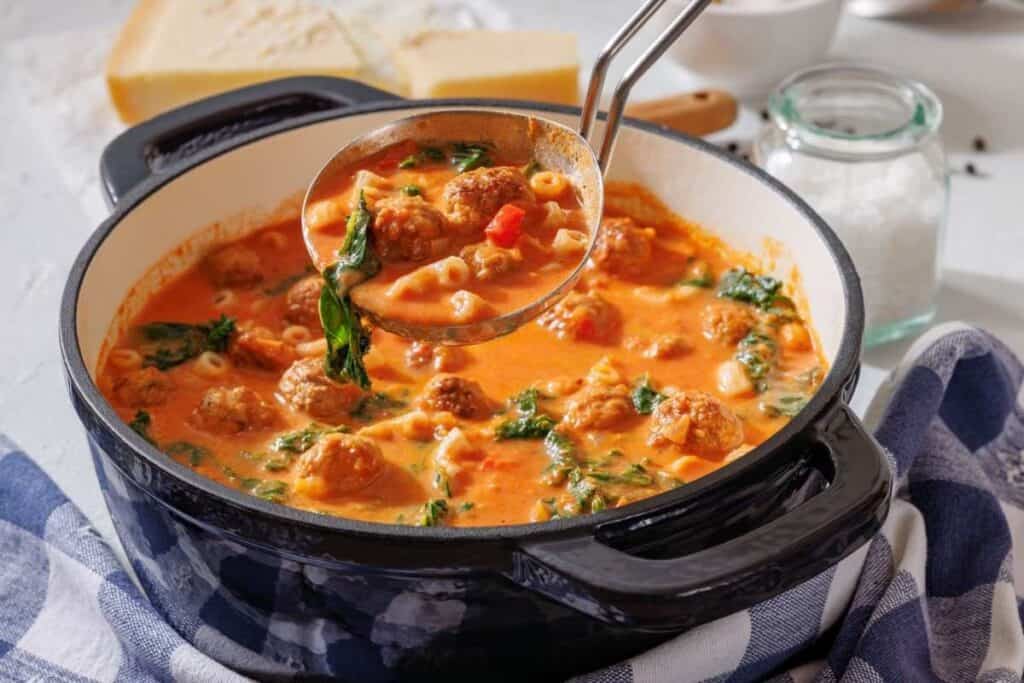A pot of tomato-based soup with meatballs, spinach, small pasta, and a ladle, placed on a blue and white cloth next to a jar of salt and a block of cheese.