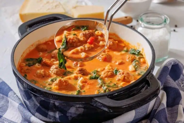 A pot of tomato-based soup with meatballs, spinach, small pasta, and a ladle, placed on a blue and white cloth next to a jar of salt and a block of cheese.