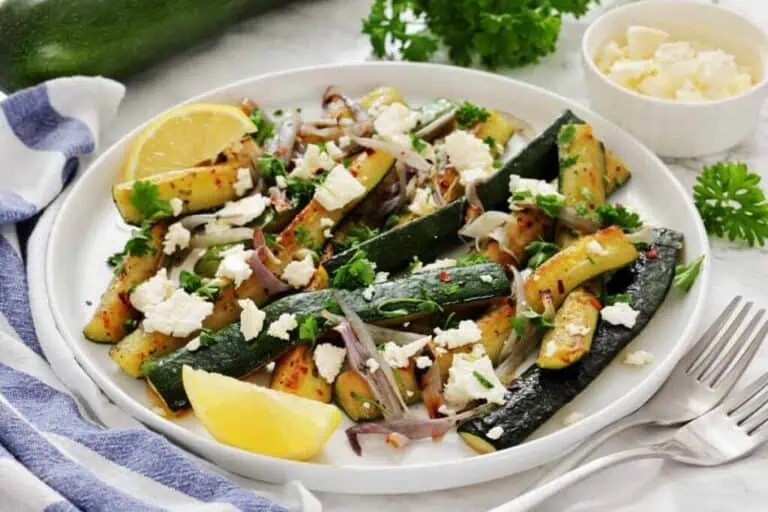 Plate of sautéed zucchini with red onions, garnished with crumbled feta cheese, parsley, and lemon wedges. A striped napkin, a fork, and extra feta are in the background.