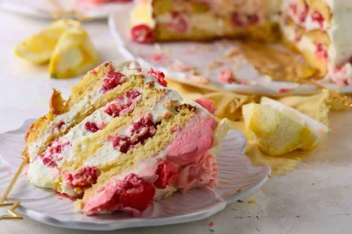 A slice of Keto Raspberry Lemon Cake with white and pink cream and pieces of fruit is on a plate, with the remaining cake and lemon wedges in the background.