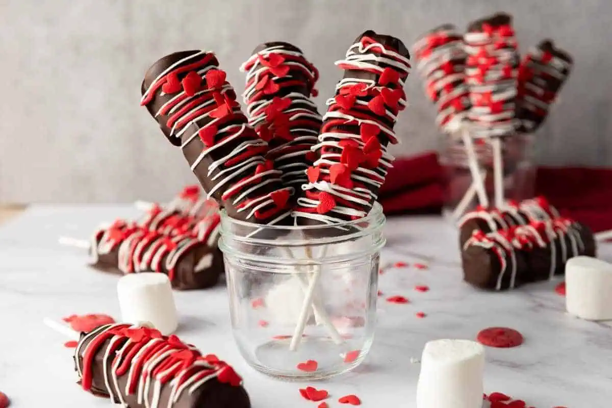 Valentine’s Day Marshmallow Pops on sticks, decorated with red heart sprinkles and white drizzle, displayed in glass jars with loose marshmallows and sprinkles around.
