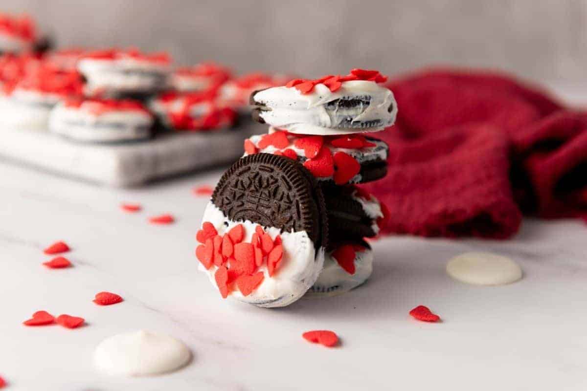 Valentine&rsquo;s Day Oreos decorated with red heart-shaped sprinkles, arranged on a white surface with a red cloth in the background.
