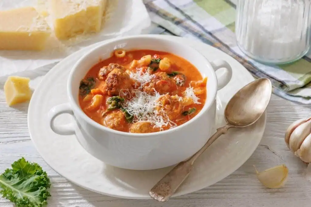 A white bowl of tomato-based soup with meatballs, pasta, greens, and grated cheese on top, placed on a white plate with a spoon beside it.