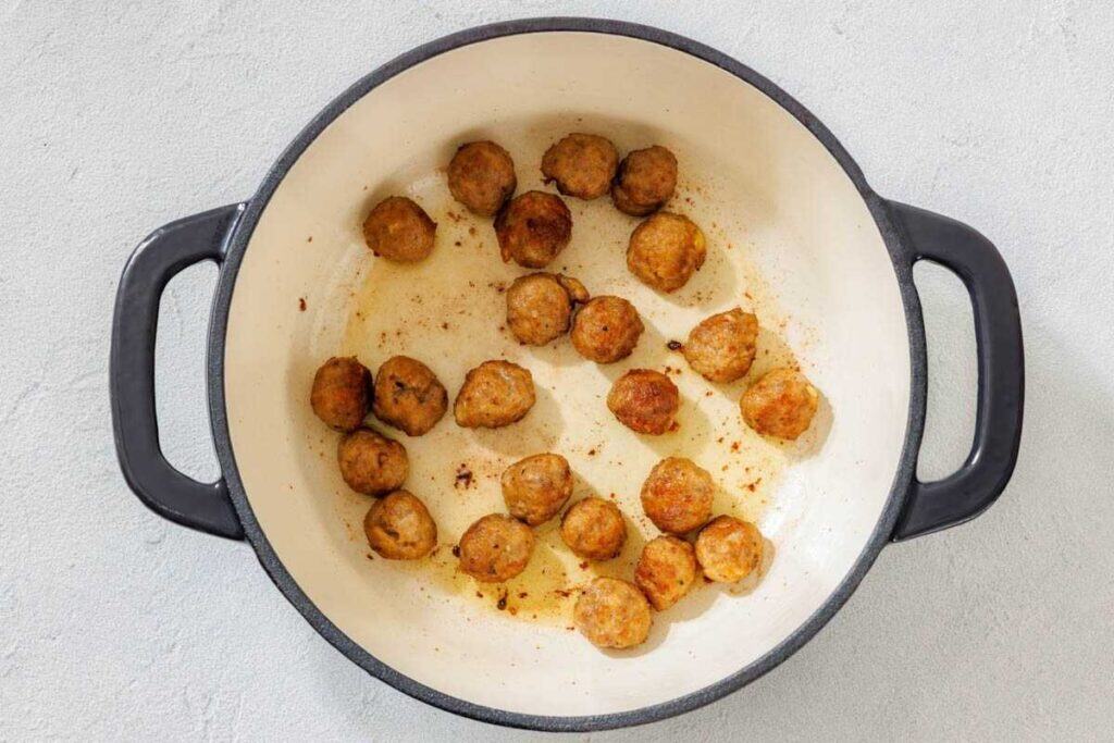 A white pot with black handles contains several browned meatballs scattered across the bottom.