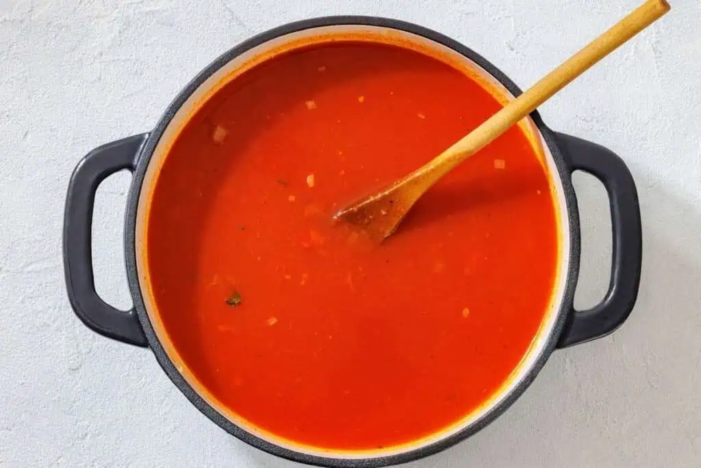 A pot of tomato soup with a wooden spoon resting inside, viewed from above on a light-colored surface.