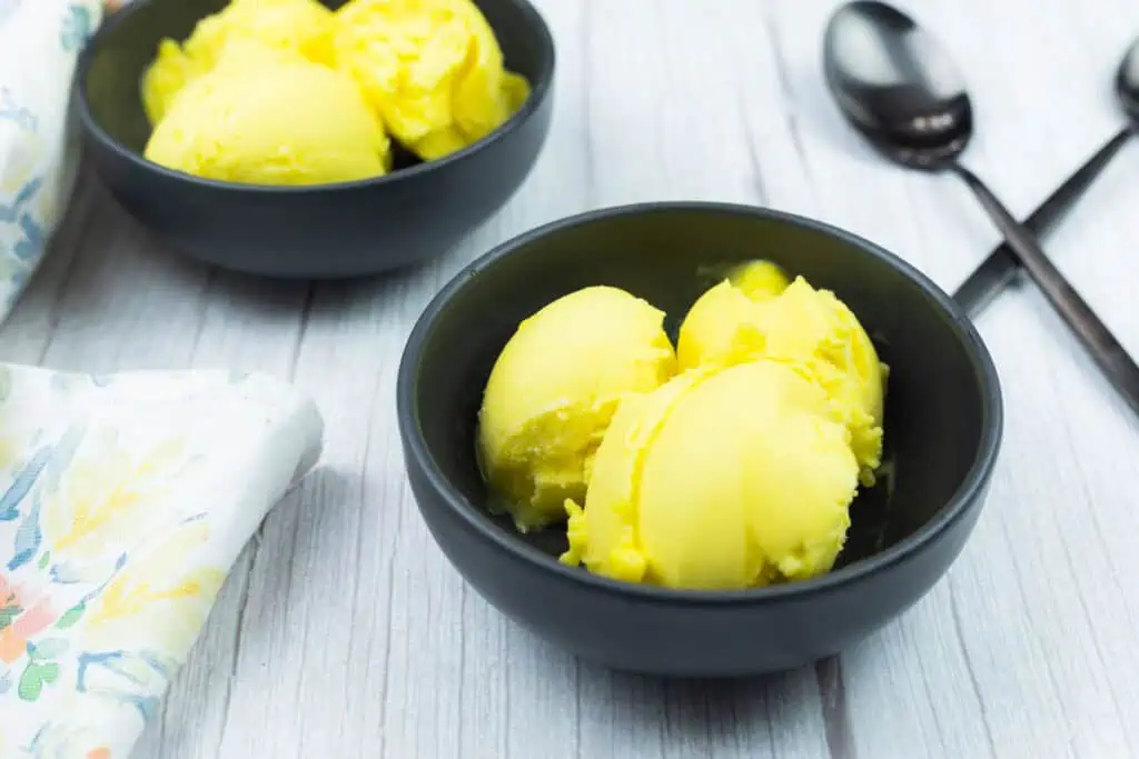 Two black bowls filled with Ninja Creami Mango Sorbet sit on a light wooden surface, with a floral napkin and two black spoons nearby.