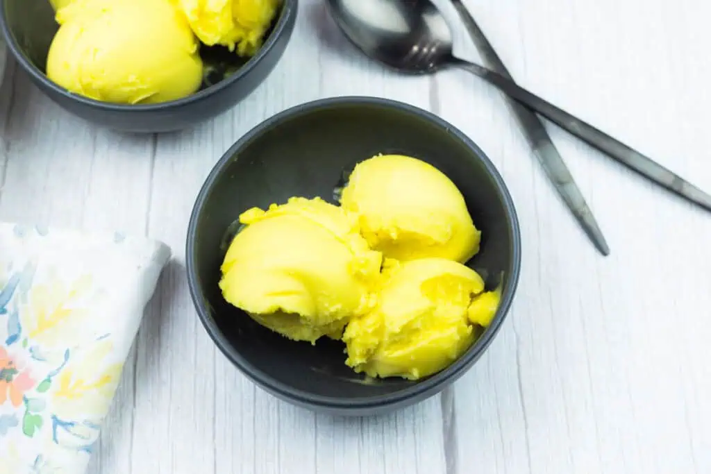 A black bowl with three scoops of yellow ice cream on a white wooden surface, next to two spoons and a floral-patterned napkin.