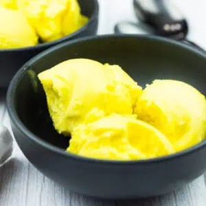 A black bowl containing three scoops of bright yellow ice cream sits on a light wooden surface, with another bowl and metal spoons in the background.