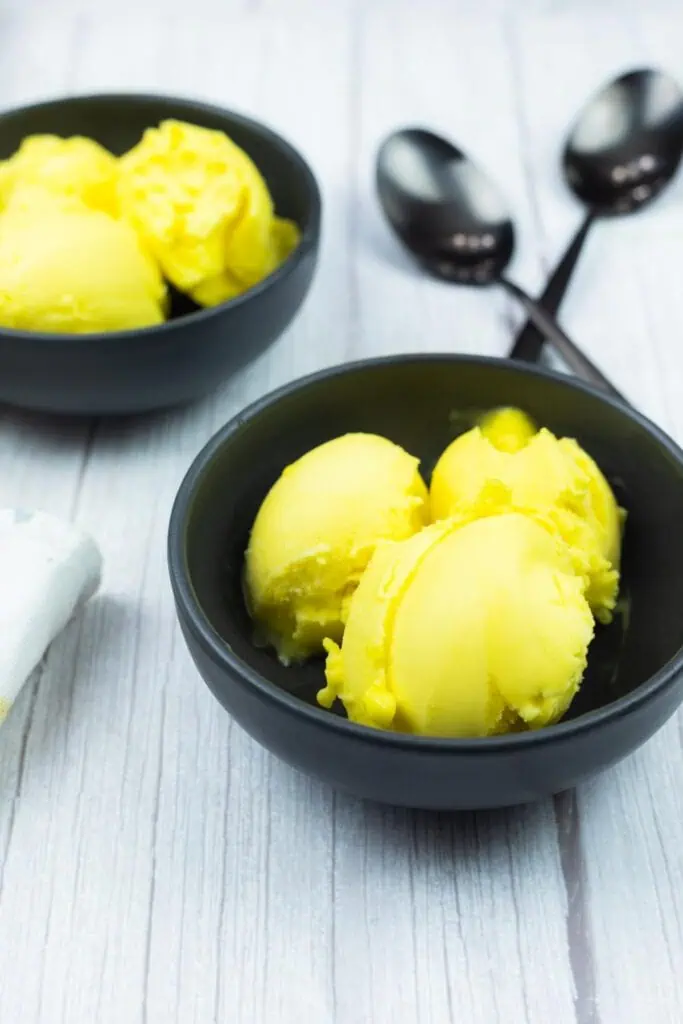 Two black bowls filled with scoops of yellow mango sorbet are on a light wooden surface, with two black spoons beside them.