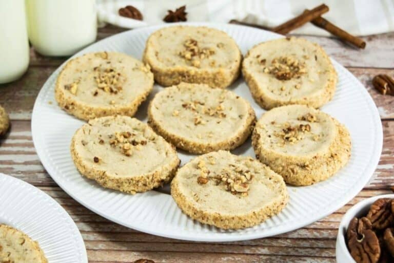 A white plate with eight round pecan shortbread cookies, some topped with chopped pecans, on a wooden table with cinnamon sticks and bottles of milk in the background.
