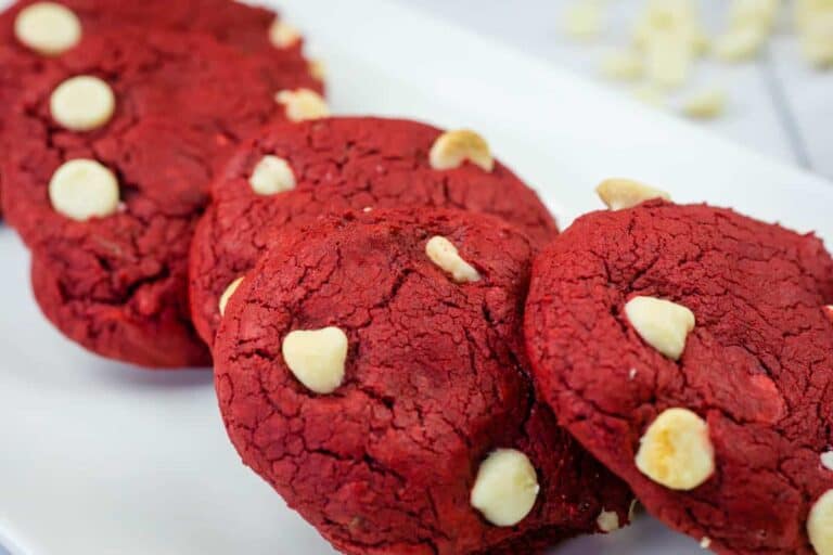 Four red cookies with white chocolate chips are arranged in a row on a white rectangular plate.