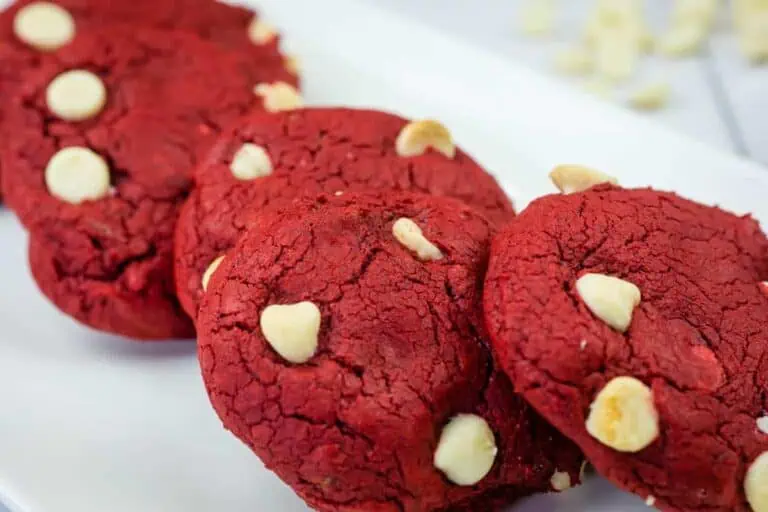 Four red cookies with white chocolate chips are arranged in a row on a white rectangular plate.