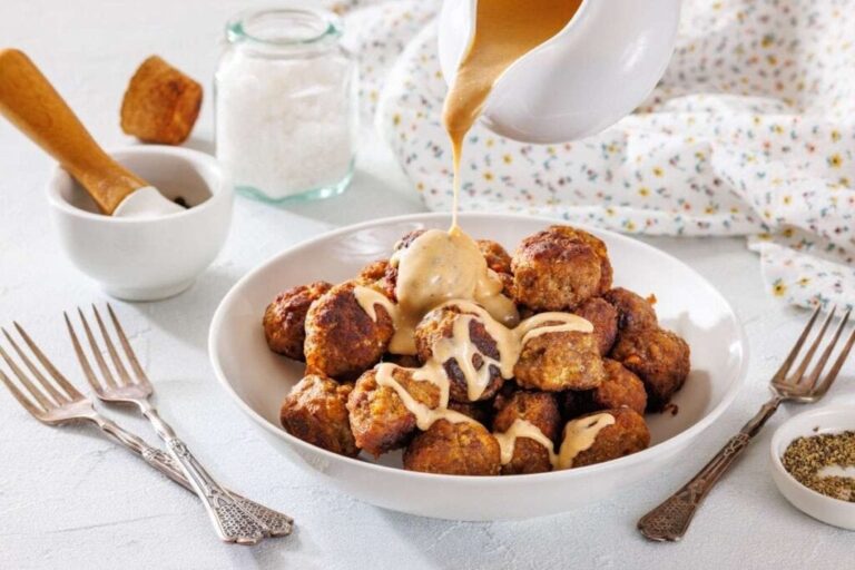 A bowl of meatballs is topped with creamy gravy being poured from a pitcher, surrounded by forks, a salt jar, and a bowl of pepper.