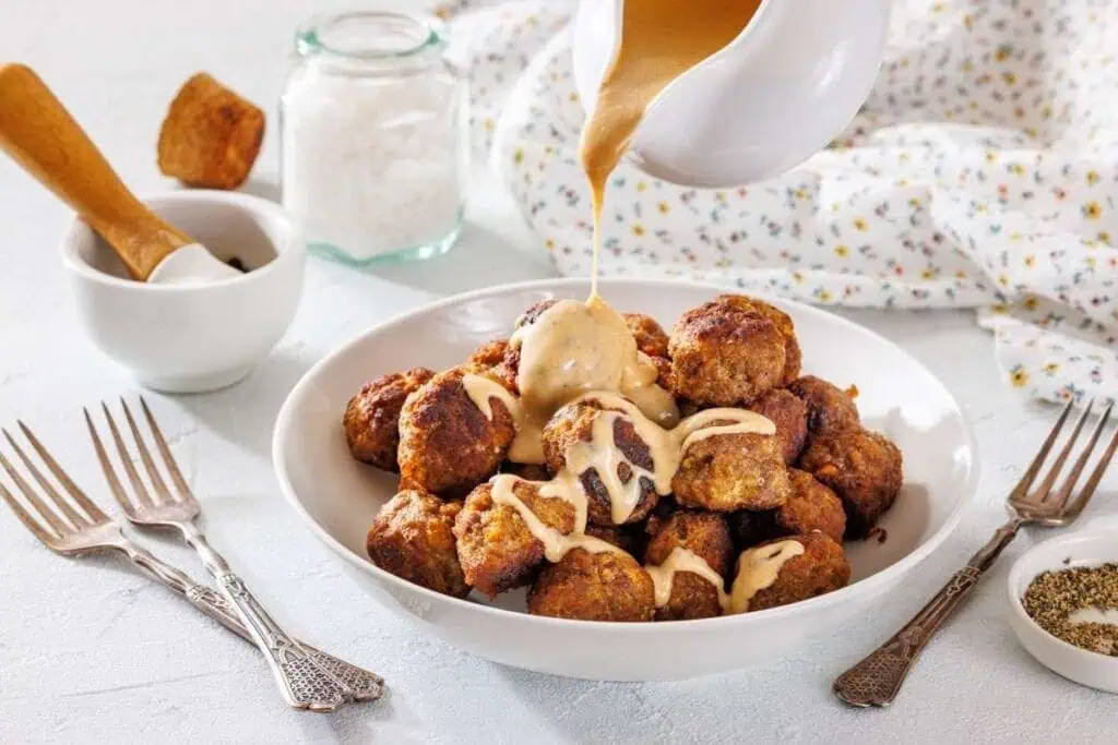 A bowl of meatballs is topped with creamy sauce being poured from a jug, surrounded by forks and seasoning jars on a white table.