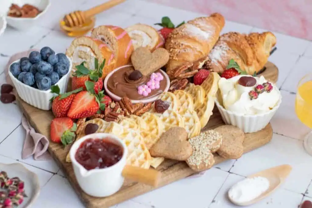 A wooden board holds croissants, waffles, strawberries, blueberries, heart-shaped cookies, jam, whipped cream, and pecans, with small bowls of spreads and garnishes on the side.