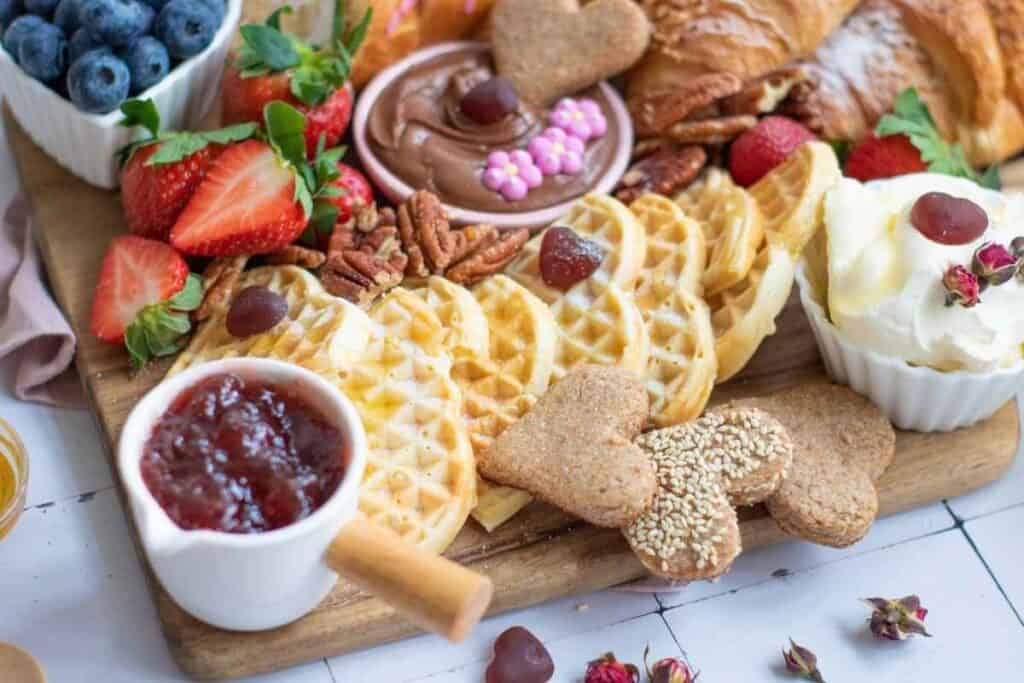 A breakfast platter with waffles, croissants, strawberries, blueberries, cookies, pecans, chocolate spread, jam, whipped cream, and garnishes on a wooden board.