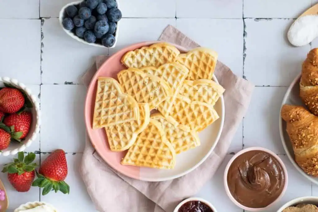 A plate of heart-shaped waffles on a pink and white plate, surrounded by bowls of blueberries, strawberries, croissants, chocolate spread, and jam on a white tiled surface.