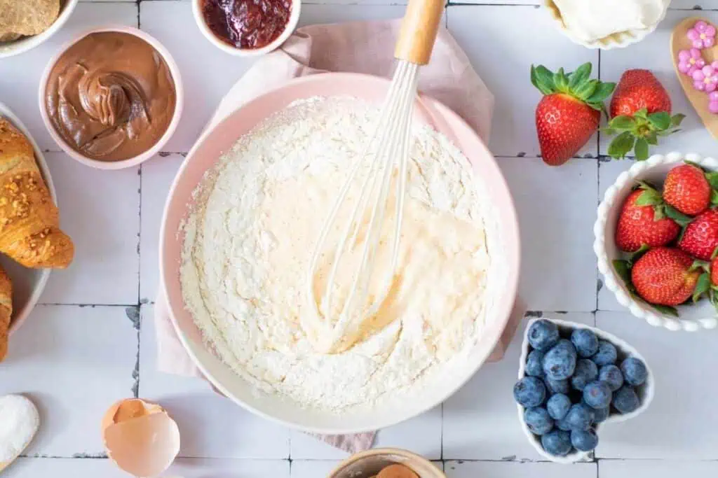 A mixing bowl with flour and a whisk surrounded by blueberries, strawberries, croissants, chocolate spread, jam, and baking ingredients on a white tiled surface.